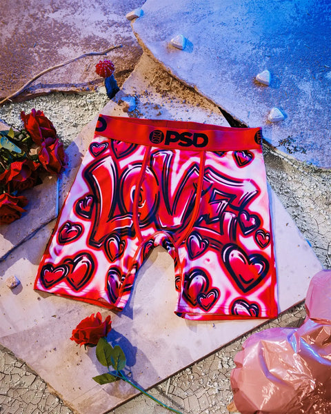 Red and pink boxer shorts with heart graphics displayed on ground surrounded by red roses and blue water.