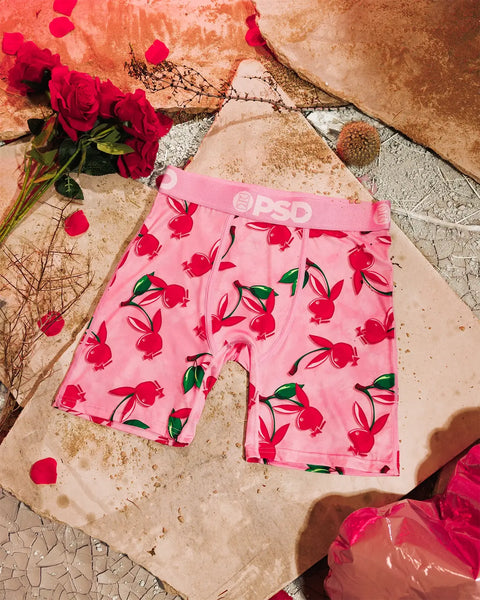 Pink boxer shorts with red cherry print and green leaves, displayed on textured beige surface with red roses and dried botani