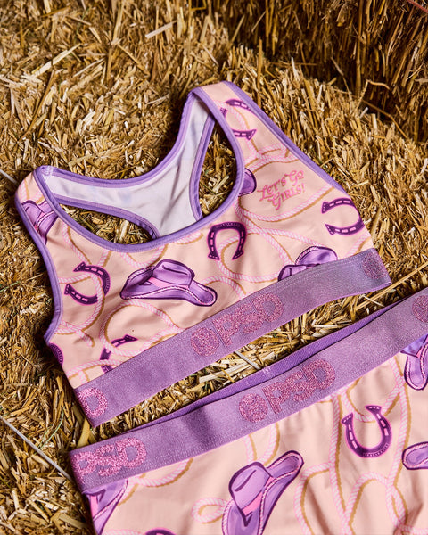 Purple and pink sports bra with cowboy hat and horseshoe print, purple trim and glitter waistband on straw background.