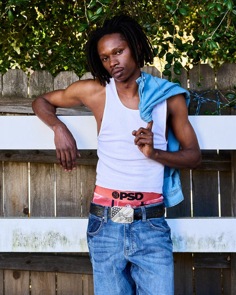 Person wearing white sleeveless tank top, blue denim shirt, jeans, and red PSD waistband against wooden fence with ivy.