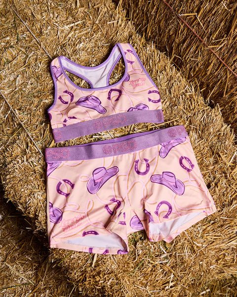 Two-piece children's swimsuit with purple cowboy hats and horseshoes on pink fabric, displayed on straw background.